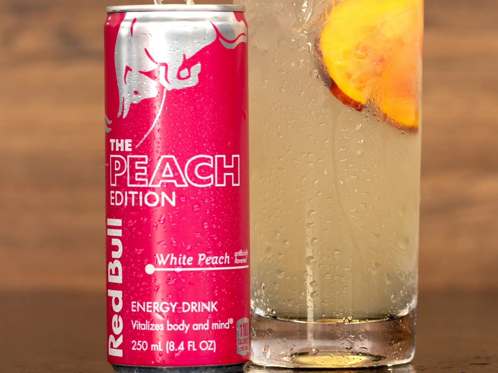 A tall White Peach & Honey Red Bull Infusion with a peach slice, beside a can of Red Bull Peach Edition