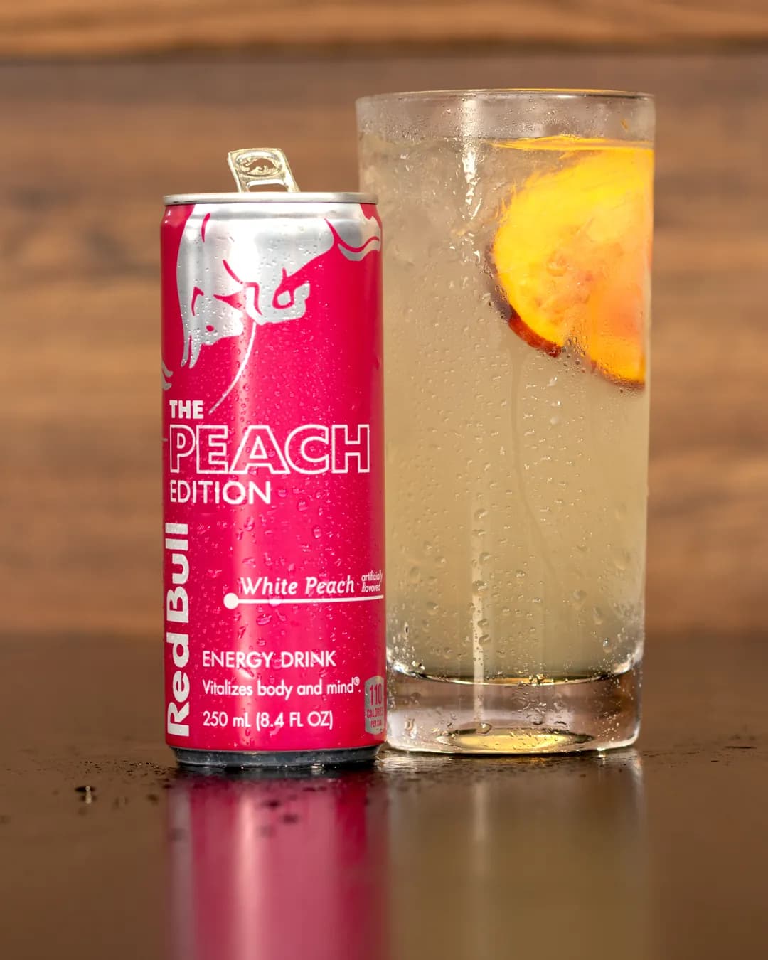 A tall White Peach & Honey Red Bull Infusion with a peach slice beside a can of Red Bull Peach Edition
